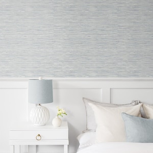 May include: A white nightstand with a drawer and a gold knob. A white ceramic lamp with a light blue lampshade sits on the nightstand. A small vase with white flowers sits next to the lamp. A white bed with a white duvet and several pillows sits in the background. The wall behind the bed is covered in a light blue and white striped wallpaper.