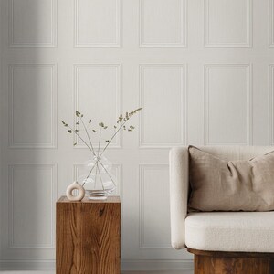 May include: A white couch with a beige pillow sits in front of a white wall with a paneled design. A wooden side table with a vase of dried flowers sits next to the couch.