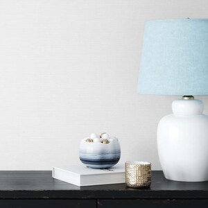 May include: A white ceramic table lamp with a light blue fabric lampshade. The lamp is on a black surface with a white book and a blue and white bowl filled with white and gold balls. A gold metal candle holder is also on the surface.