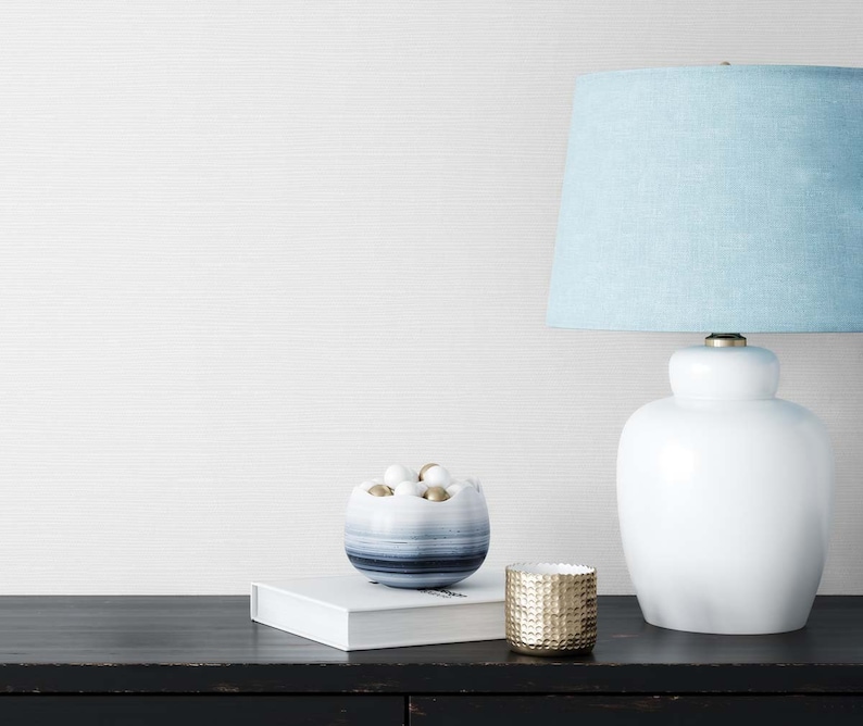 May include: A white ceramic table lamp with a light blue fabric lampshade. The lamp is on a black surface with a white book and a blue and white bowl filled with white and gold balls. A gold metal candle holder is also on the surface.