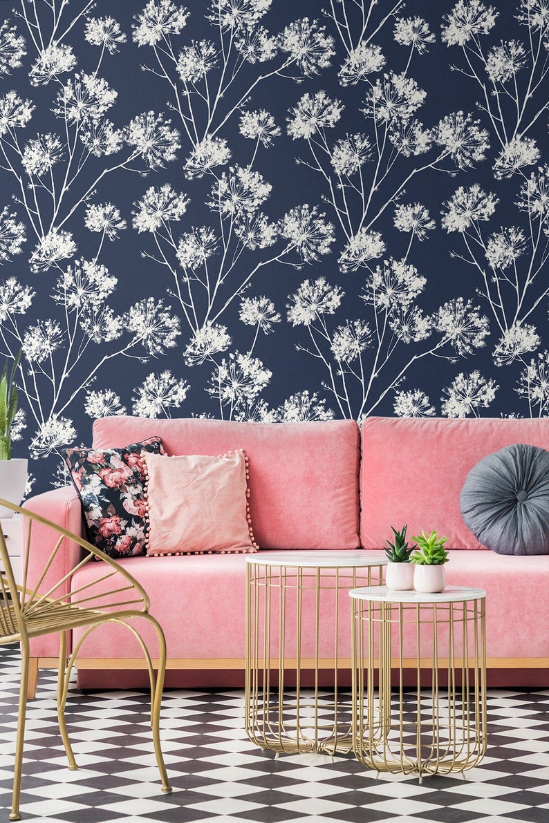 May include: A pink sofa with floral patterned pillows and a gray throw pillow sits in front of a wall with a white floral pattern on a dark blue background. Two gold wire-frame side tables with white marble tops are in front of the sofa. One table has two small potted plants on it.