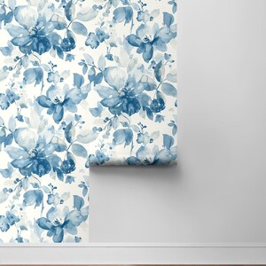 May include: A roll of blue and white floral wallpaper with a watercolor design. The wallpaper is being unrolled on a white wall.