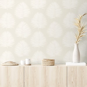 May include: A beige wall with a white palm leaf pattern. A wooden shelf with a woven basket, two candles, and a small woven mat. A vase with dried beige branches sits on a white book on the shelf.