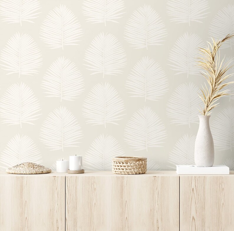 May include: A beige wall with a white palm leaf pattern. A wooden shelf with a woven basket, two candles, and a small woven mat. A vase with dried beige branches sits on a white book on the shelf.
