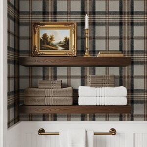 May include: A bathroom scene with a plaid wallpaper background. Two wooden shelves hold folded towels in shades of brown and white. A framed landscape painting and a brass candlestick with a lit candle sit on the top shelf. A brass towel rack with white towels hangs below.