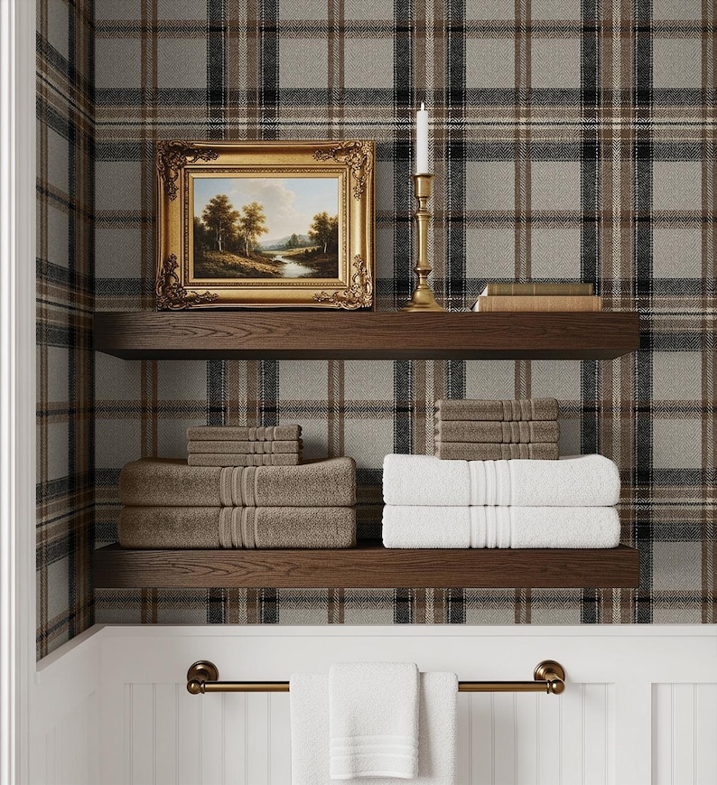 May include: A bathroom scene with a plaid wallpaper background. Two wooden shelves hold folded towels in shades of brown and white. A framed landscape painting and a brass candlestick with a lit candle sit on the top shelf. A brass towel rack with white towels hangs below.