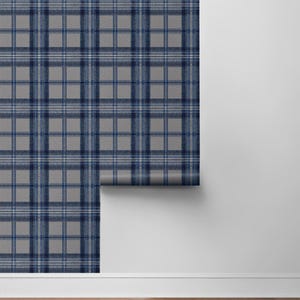 May include: A roll of gray and navy blue plaid wallpaper. The wallpaper features a classic tartan pattern with intersecting lines in varying shades of blue and white. The roll is partially unrolled, revealing the pattern against a white wall and wooden floor.