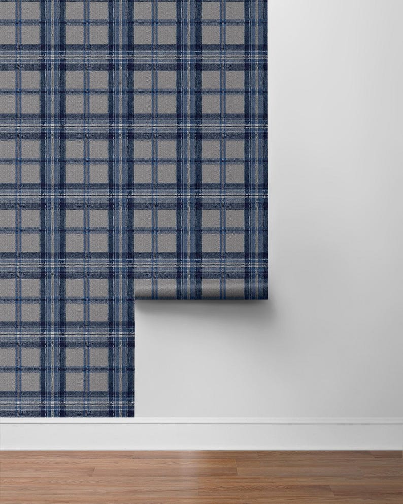 May include: A roll of gray and navy blue plaid wallpaper. The wallpaper features a classic tartan pattern with intersecting lines in varying shades of blue and white. The roll is partially unrolled, revealing the pattern against a white wall and wooden floor.