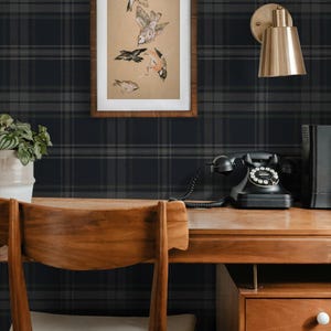 May include: A black and gray plaid wallpaper with a framed print of birds and a gold wall sconce. A wooden desk with a white chair, a black rotary phone, and a wooden drawer unit with two drawers.