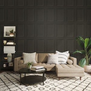 May include: A sectional sofa with a light brown leather upholstery and a dark brown wood frame. The sofa is positioned in front of a wall with a dark gray wallpaper featuring a paneled design. The sofa has two pillows with a white and gray geometric pattern and one pillow with a white and gray striped pattern. A black coffee table with two tiers sits in front of the sofa. A potted plant sits on the coffee table. A small potted plant sits on a shelf unit to the left of the sofa. A large potted plant sits to the right of the sofa.