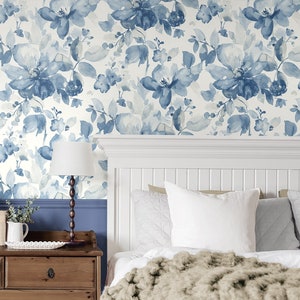 May include: A white bedroom with a blue floral wallpaper. The bed has a white headboard and a white duvet with a brown throw blanket. There are white pillows on the bed and a lamp on a wooden nightstand.