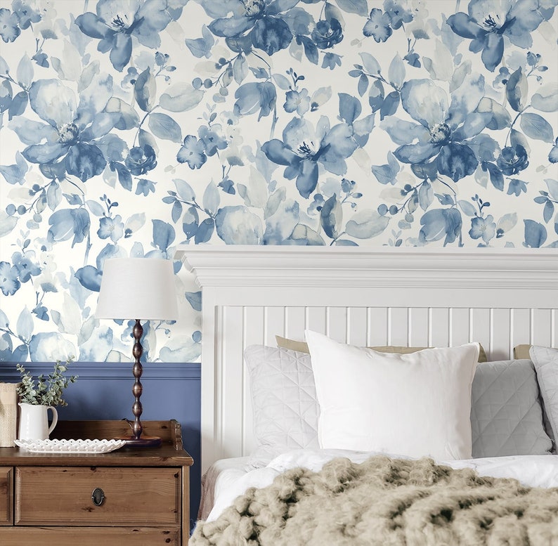 May include: A white bedroom with a blue floral wallpaper. The bed has a white headboard and a white duvet with a brown throw blanket. There are white pillows on the bed and a lamp on a wooden nightstand.