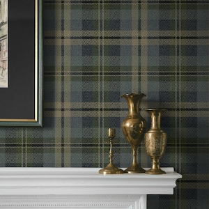 May include: A framed watercolor painting of a European street scene hangs above a white mantel. Three antique brass vases and a candlestick holder are arranged on the mantel. The background is a plaid wallpaper in shades of green, blue, and tan.