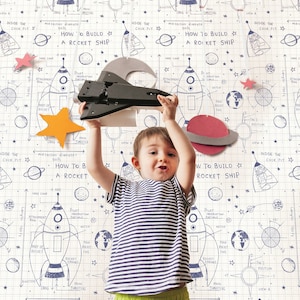 May include: A young child holds a toy space shuttle in front of a white wall with a repeating pattern of rocket ships, planets, and stars. The pattern includes the text "How to Build a Rocket Ship".