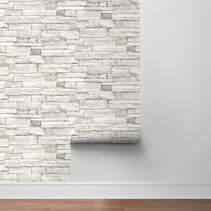 May include: A roll of white brick peel and stick wallpaper is partially unrolled on a white wall with white baseboards and a wood floor.