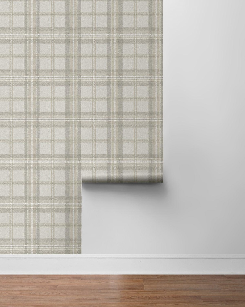 May include: A roll of wallpaper with a neutral plaid pattern in shades of beige, tan, and white. The wallpaper is partially unrolled, revealing the pattern against a white wall and hardwood floor.