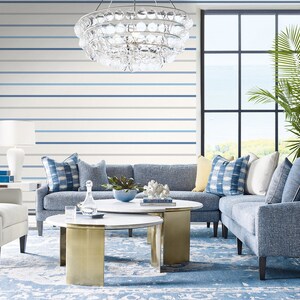 Puede incluir: Un sal&oacute;n moderno con una combinaci&oacute;n de colores azul y blanco. La habitaci&oacute;n cuenta con un sof&aacute; azul con cojines estampados, un sill&oacute;n blanco y dos mesas de centro redondas con bases doradas. Una gran ventana ofrece vistas al mar.