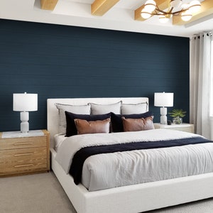 Op de afbeelding: Een wit gestoffeerd bed met een grijs en zwart gestreept dekbed. Het bed staat in een slaapkamer met een donkerblauwe accentmuur, houten balken aan het plafond en twee witte lampen op nachtkastjes.