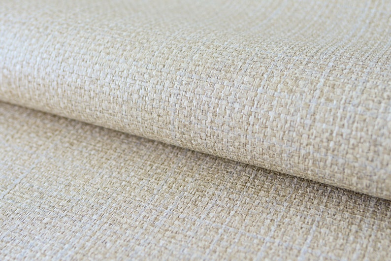 May include: Close-up of a beige woven fabric with a textured surface. The fabric is folded, revealing the weave pattern and subtle variations in color. The material appears to be a natural fiber.