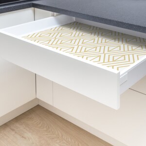 K&ouml;nnte beinhalten: Eine wei&szlig;e K&uuml;chenschublade ist teilweise ge&ouml;ffnet und zeigt eine geometrisch gemusterte Auskleidung. Die Auskleidung weist ein sich wiederholendes Design aus goldfarbenen Linien auf wei&szlig;em Hintergrund auf. Die Schublade befindet sich vor einer schwarzen Arbeitsplatte.
