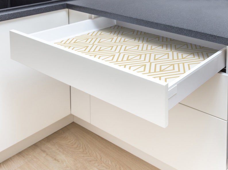 K&ouml;nnte beinhalten: Eine wei&szlig;e K&uuml;chenschublade ist teilweise ge&ouml;ffnet und zeigt eine geometrisch gemusterte Auskleidung. Die Auskleidung weist ein sich wiederholendes Design aus goldfarbenen Linien auf wei&szlig;em Hintergrund auf. Die Schublade befindet sich vor einer schwarzen Arbeitsplatte.
