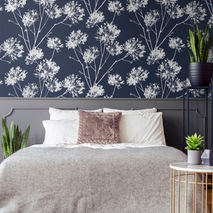 May include: A gray bed with white bedding and a brown throw pillow. The headboard is gray and has a floral wallpaper behind it. There is a gold side table with a white top and a green plant on it.