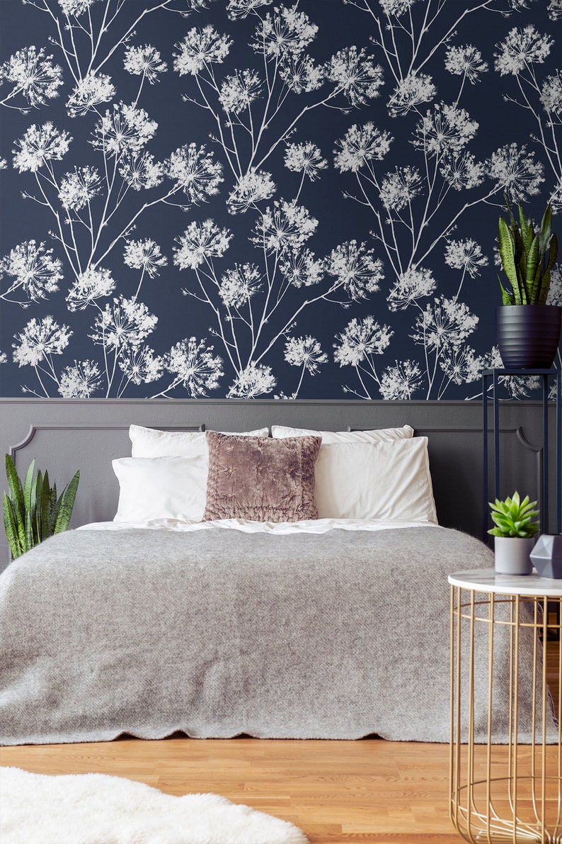 May include: A gray bed with white bedding and a brown throw pillow. The headboard is gray and has a floral wallpaper behind it. There is a gold side table with a white top and a green plant on it.