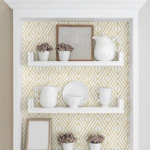 K&ouml;nnte beinhalten: Wei&szlig;es, in die Wand eingelassenes Regal mit geometrischem Tapetenhintergrund. Die Regale zeigen wei&szlig;e Teetassen, eine Teekanne, einen Bilderrahmen und kleine Topfpflanzen. Die Tapete hat ein gold-wei&szlig;es Diamantenmuster.