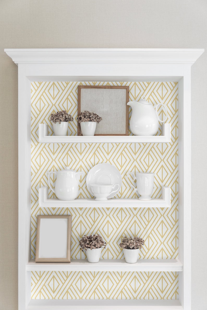 K&ouml;nnte beinhalten: Wei&szlig;es, in die Wand eingelassenes Regal mit geometrischem Tapetenhintergrund. Die Regale zeigen wei&szlig;e Teetassen, eine Teekanne, einen Bilderrahmen und kleine Topfpflanzen. Die Tapete hat ein gold-wei&szlig;es Diamantenmuster.