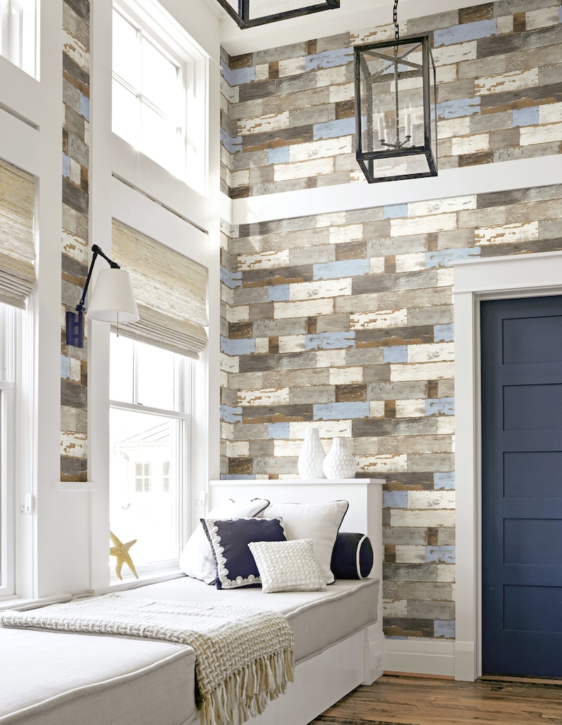 May include: A white window seat with a light brown blanket and blue and white pillows in front of a wall with a wood plank wallpaper design. A black door is on the right side of the image.
