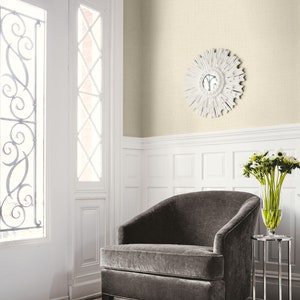 May include: A room interior featuring a gray velvet armchair, a small silver side table with a vase of flowers, and a white sunburst mirror. The walls are a light beige color, and the floor has a fluffy white rug.
