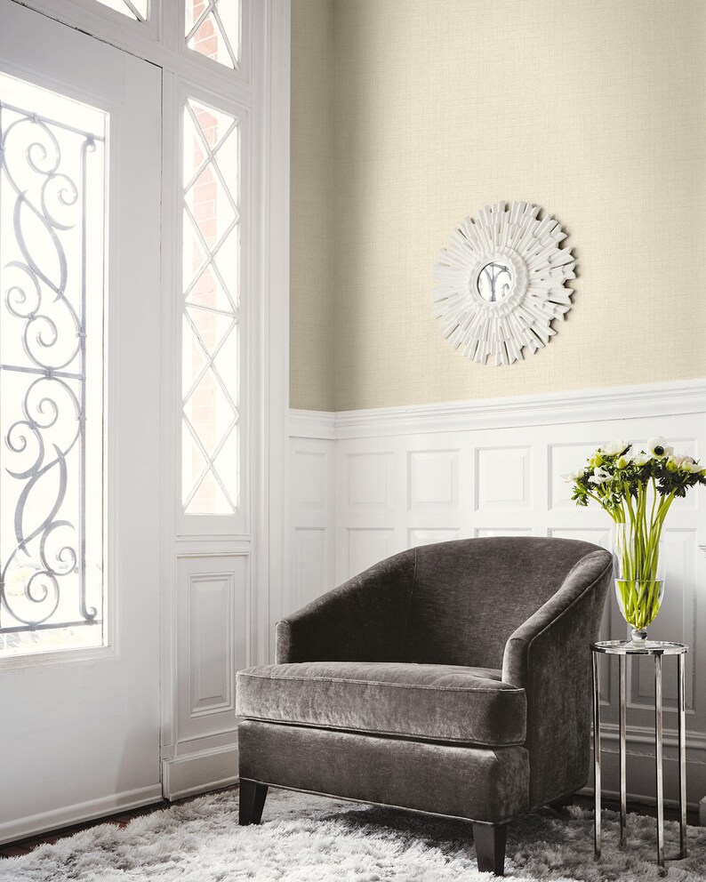 May include: A room interior featuring a gray velvet armchair, a small silver side table with a vase of flowers, and a white sunburst mirror. The walls are a light beige color, and the floor has a fluffy white rug.