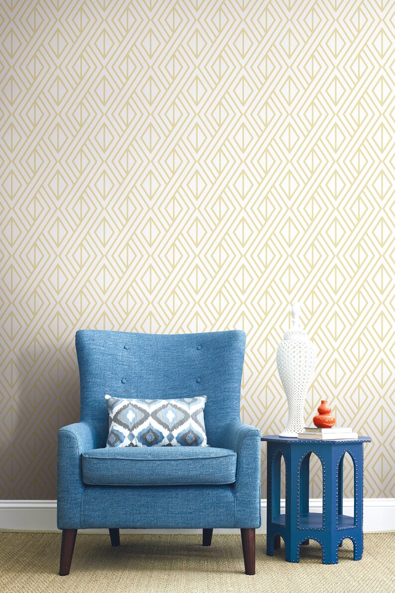 K&ouml;nnte beinhalten: Ein blauer Sessel mit einem gemusterten Kissen steht vor einer Wand mit einem geometrischen Goldmuster. Ein kleiner blauer Beistelltisch tr&auml;gt eine wei&szlig;e Vase und ein orangefarbenes Dekorationsobjekt. Der Raum hat eine neutrale Farbpalette.