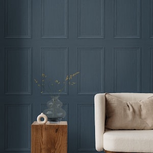 May include: A white armchair with a beige cushion sits in front of a wall with a dark blue, wood-paneled wallpaper. A wooden side table with a clear glass vase and a small white ceramic sculpture sits in front of the chair.