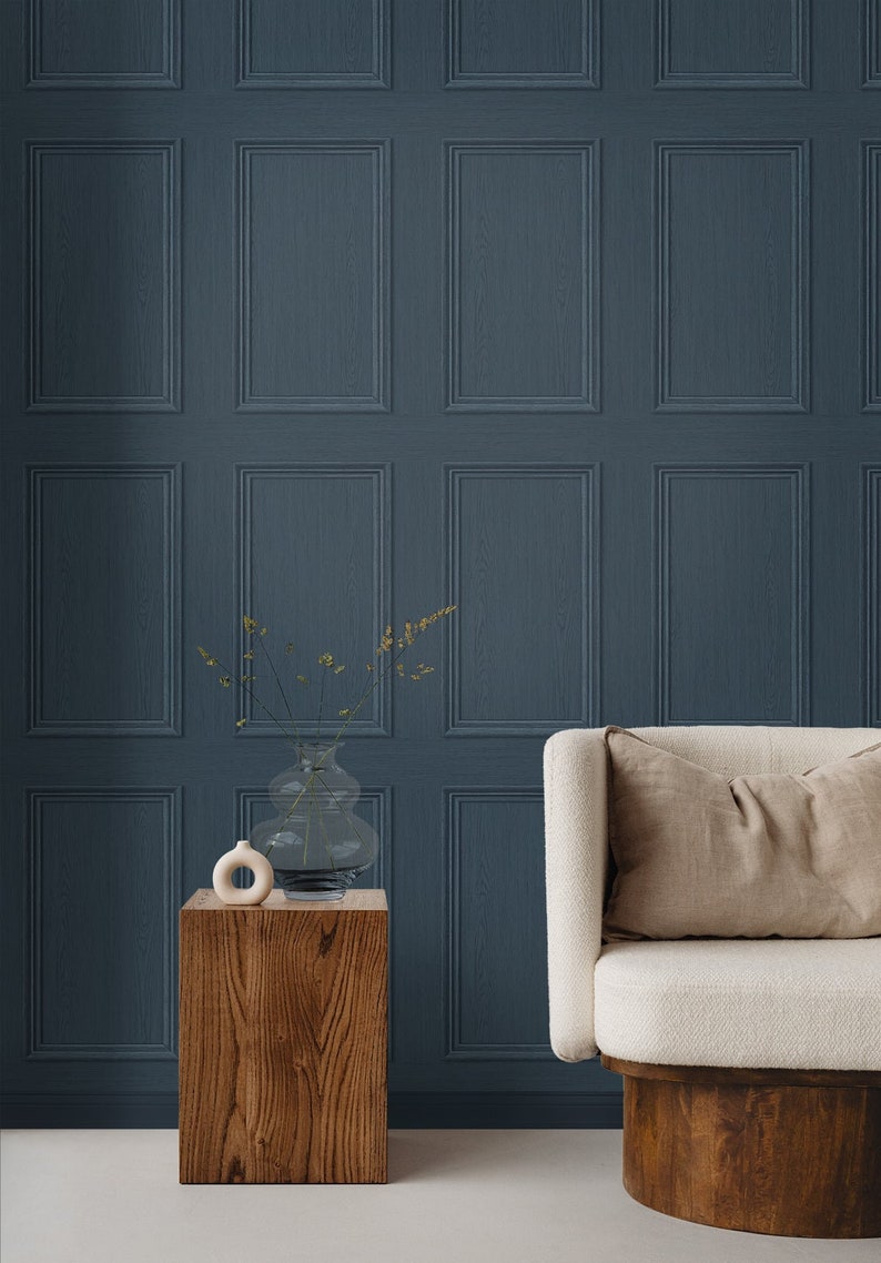 May include: A white armchair with a beige cushion sits in front of a wall with a dark blue, wood-paneled wallpaper. A wooden side table with a clear glass vase and a small white ceramic sculpture sits in front of the chair.