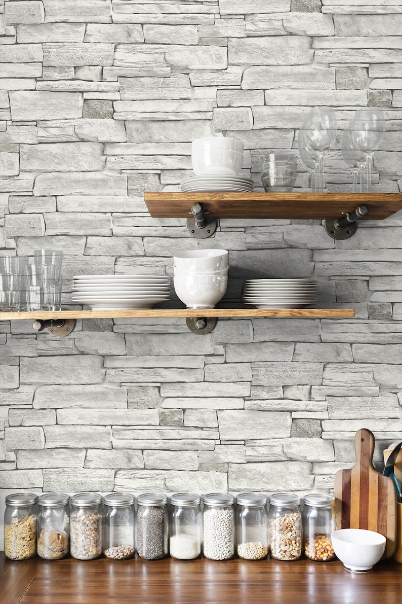 May include: A white brick wall with two wooden shelves holding various dishes and glasses. Below the shelves are ten glass jars filled with different types of grains and beans. A white bowl and a wooden cutting board are on the counter.