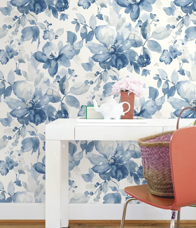 May include: A white desk with a blue and white floral wallpaper background. A white teapot, a small green box, and a pink flower arrangement are on the desk. A red chair with a woven basket is in the foreground.