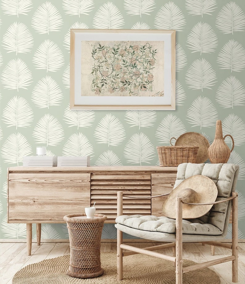May include: A light green wallpaper with a white palm leaf pattern. A wooden dresser with a framed print of a floral design sits on top of the dresser. A wicker basket, a vase, and a chair with a straw hat are in the background.