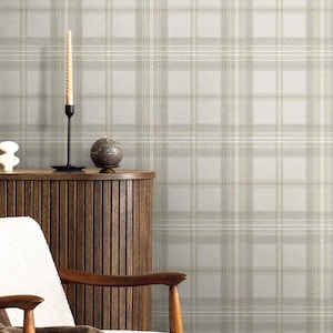 May include: A neutral-toned interior scene featuring a plaid wallpaper backdrop. A wooden cabinet, a white chair with wooden arms, and a brown throw blanket are in the foreground. A tall, spiral candle and a round candle sit on the cabinet.
