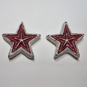 Puede incluir: Cuatro objetos decorativos en forma de estrella. Cada estrella tiene un centro rojo con un diseño de estrella blanca y un borde blanco. Las estrellas están dispuestas en fila sobre una superficie blanca. Las estrellas miden unos 5 cm de diámetro.