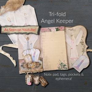 May include: A tri-fold journal with a cream-colored paper cover and a decorative angel design. The journal has a note pad, tags, pockets, and ephemera. The text "As Seen on Youtube!" and "Tri-fold Angel Keeper" is visible on the cover.