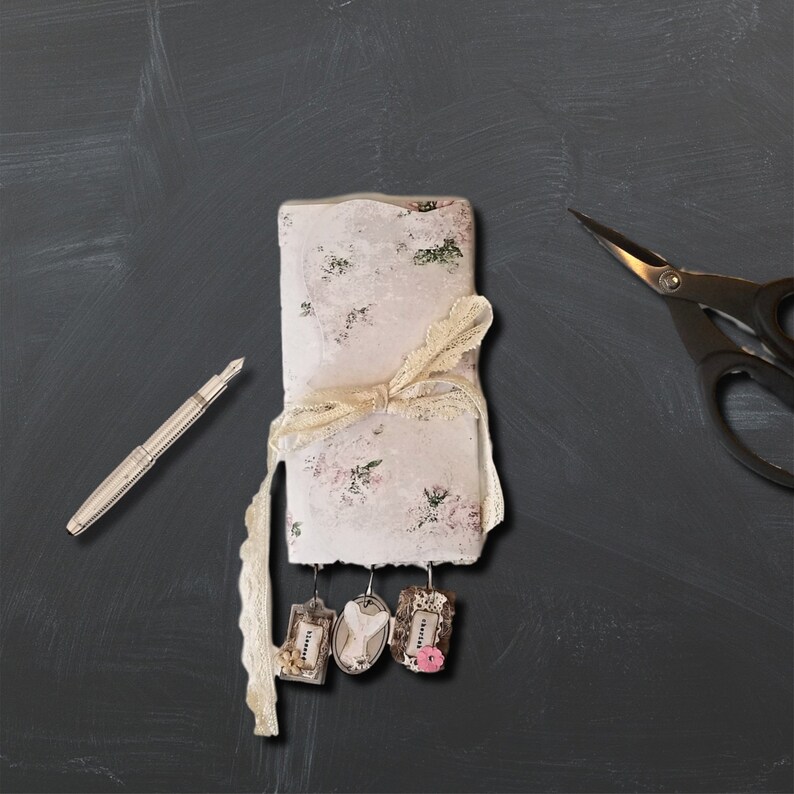 May include: A white journal with a floral pattern and a delicate lace ribbon tied around it. The journal has three small tags hanging from it, each with a different design.