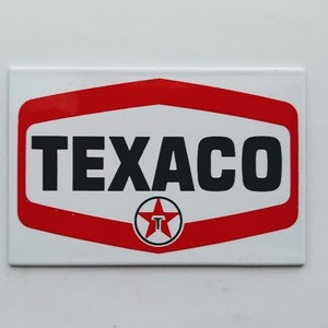 May include: Rectangular white Texaco sign with a red and white border. The word "TEXACO" is in bold black letters. A red star with a white "T" inside is at the bottom. The sign is a vintage style.