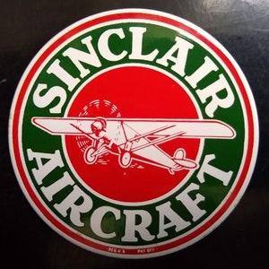 May include: A round, green and red sign with a white airplane in the center. The sign says "Sinclair Aircraft" in a white circle around the airplane. The text "Reg. U.S. Pat. Off." is at the bottom of the sign.