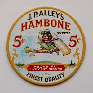 May include: Round vintage advertisement for J.P. Alley's Hambone Sweets and cigars. The image features a cartoon character in a biplane with the text "GOING OVER". The text "5¢" appears twice, with the words "FINEST QUALITY" at the bottom.