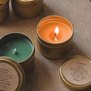 Op de afbeelding: Een verzameling van zes kleine, gouden metalen blikken met deksels. Elk blik bevat een sojawas kaars. De kaarsen zijn gelabeld met de namen "Citrus Sun", "Duende" en "Citrus Sun".