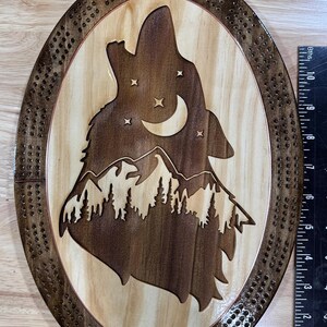 May include: Wooden cribbage board with an oval shape, featuring a howling wolf silhouette against a mountain and starry night scene. The board is made of light and dark brown wood, with a decorative border and drilled holes for pegs.