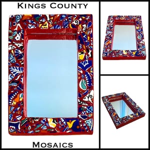 May include: A rectangular mirror with a red frame, adorned with a mosaic of colorful ceramic tiles. The mosaic features blue, yellow, and white patterns. The text "Kings County Mosaics" is visible.
