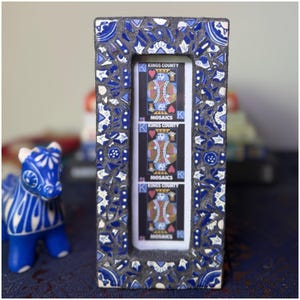 May include: A rectangular mosaic frame with a blue and white floral pattern surrounds three playing cards featuring the King of Hearts. The frame's border is dark gray. The cards read "Kings County Mosaics."