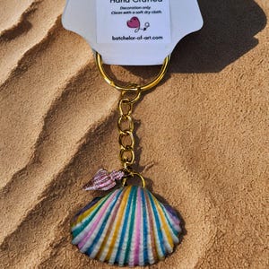 May include: A handcrafted seashell keyring with a gold-coloured chain and ring. The seashell is painted with stripes of blue, yellow, pink, and white. A smaller pink seashell charm hangs above the larger one. The tag reads "Hand Crafted" and "batchelor-of-art.com".